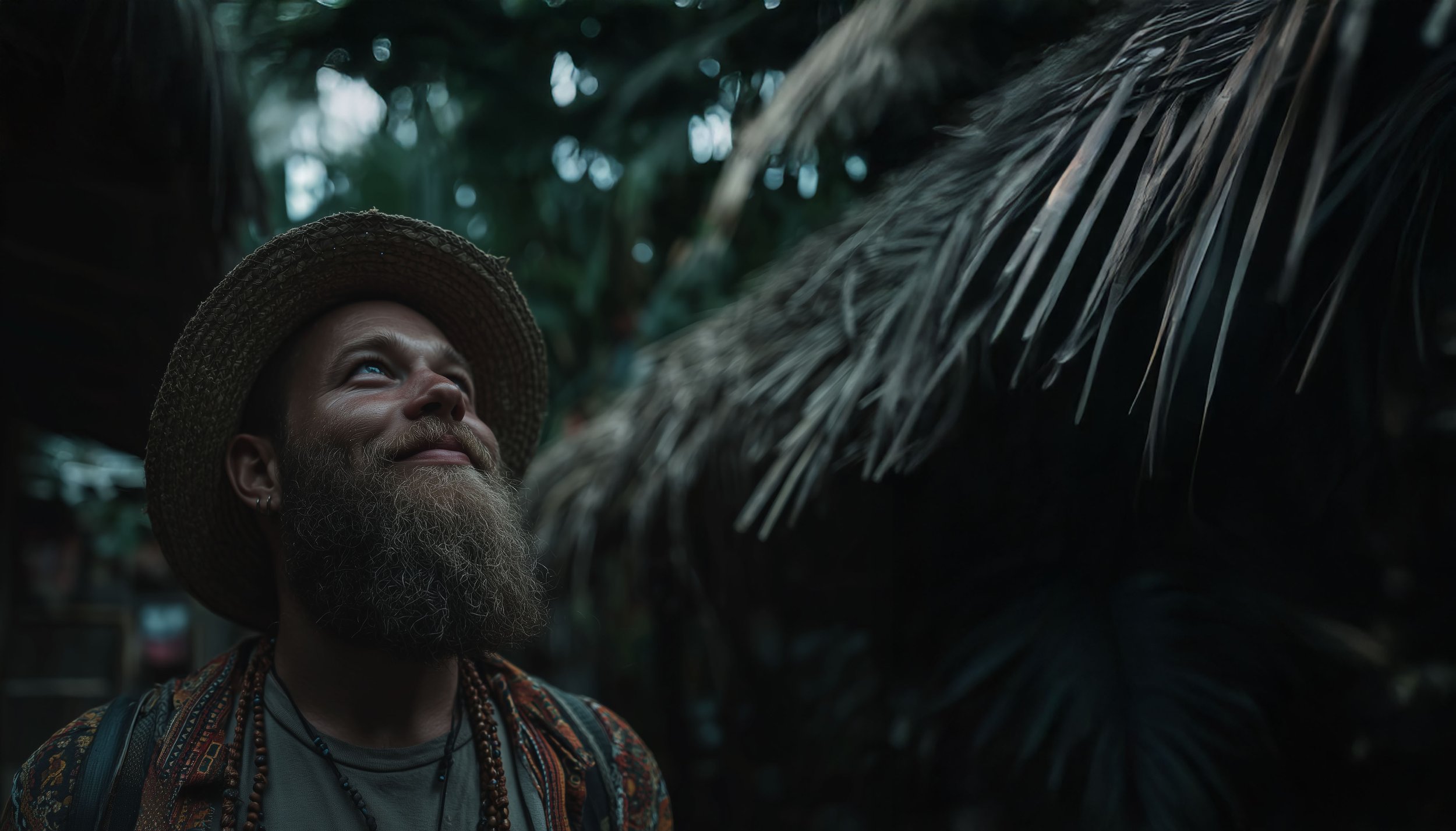 A smiling man with a beard wearing a brown cap, denim jacket, and hoodie outdoors among green plants and trees